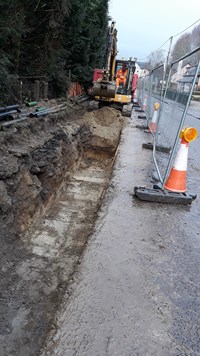 A trench next to a road, a construction vehicle with an excavation attachment is present at the end of the trench.