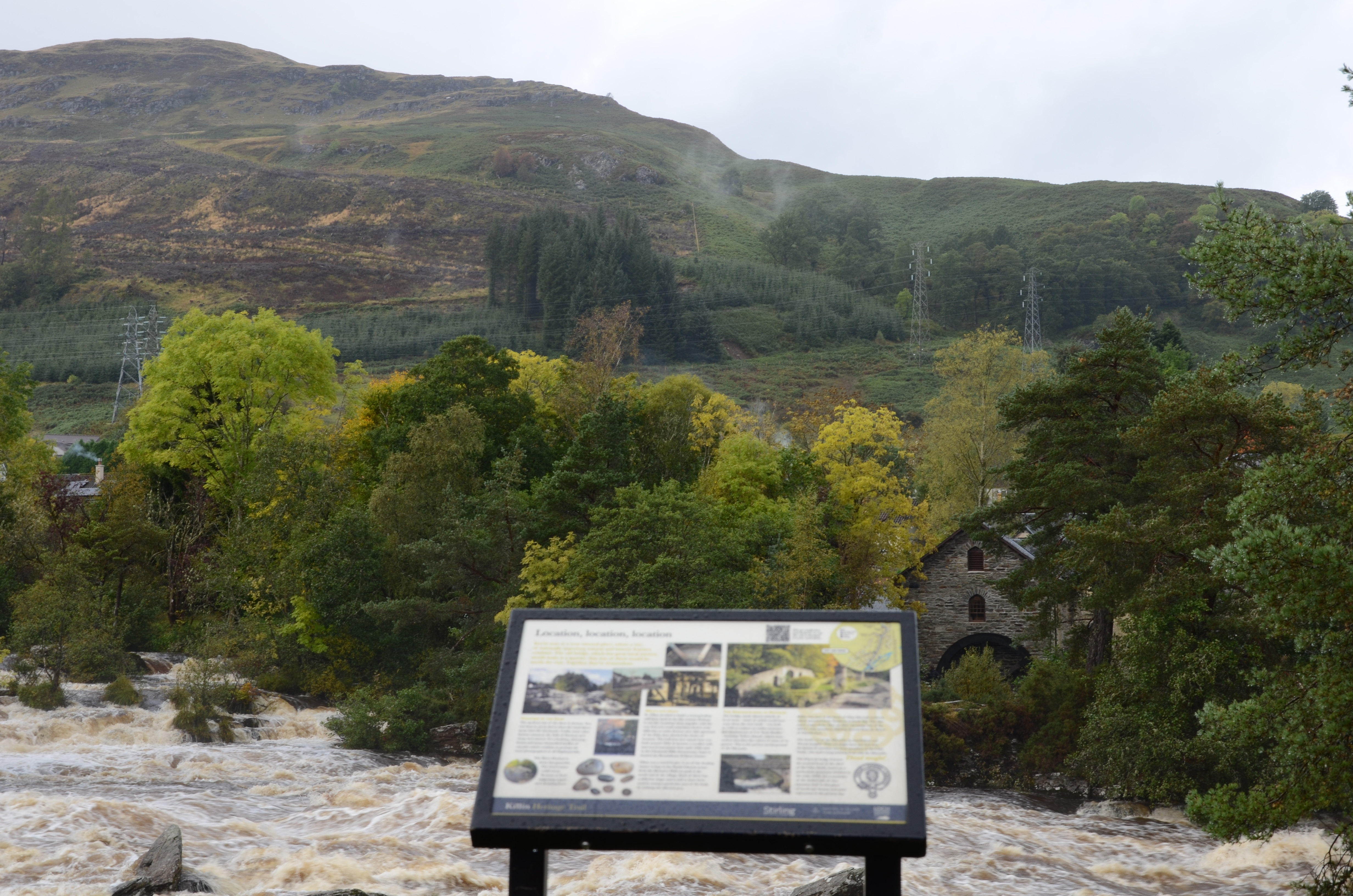 View from an out of focus information board across an area of agitated water up to a hillside where an overhead line of metal transmission towers are present.