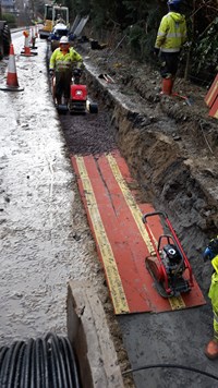 Contractors in PPE with machinery in a trench by a road.