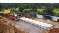 Low concrete structures in an excavated area near a river. A flattening construction vehicle is parked nearby.