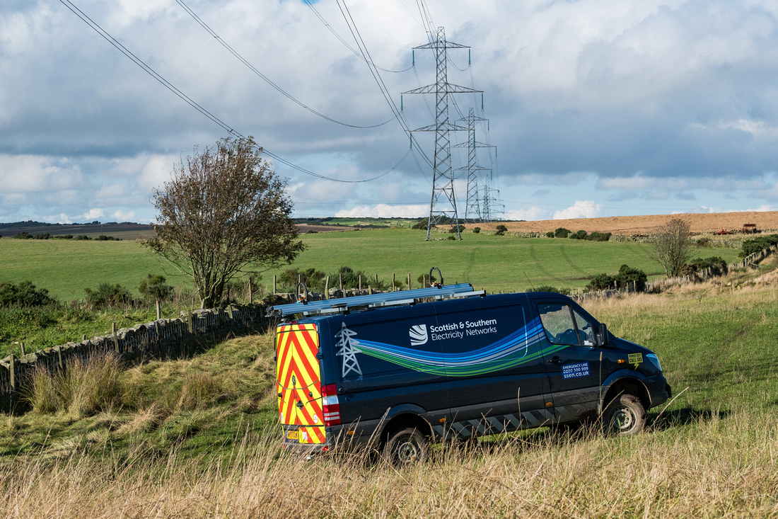 An SSEN Transmission vehicle parked off-road. In the background is a series of metal transmission tower forming an overhead line.