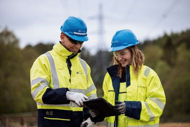 Two people in safety gear reviewing information on a tablet at an outdoor transmission work site.