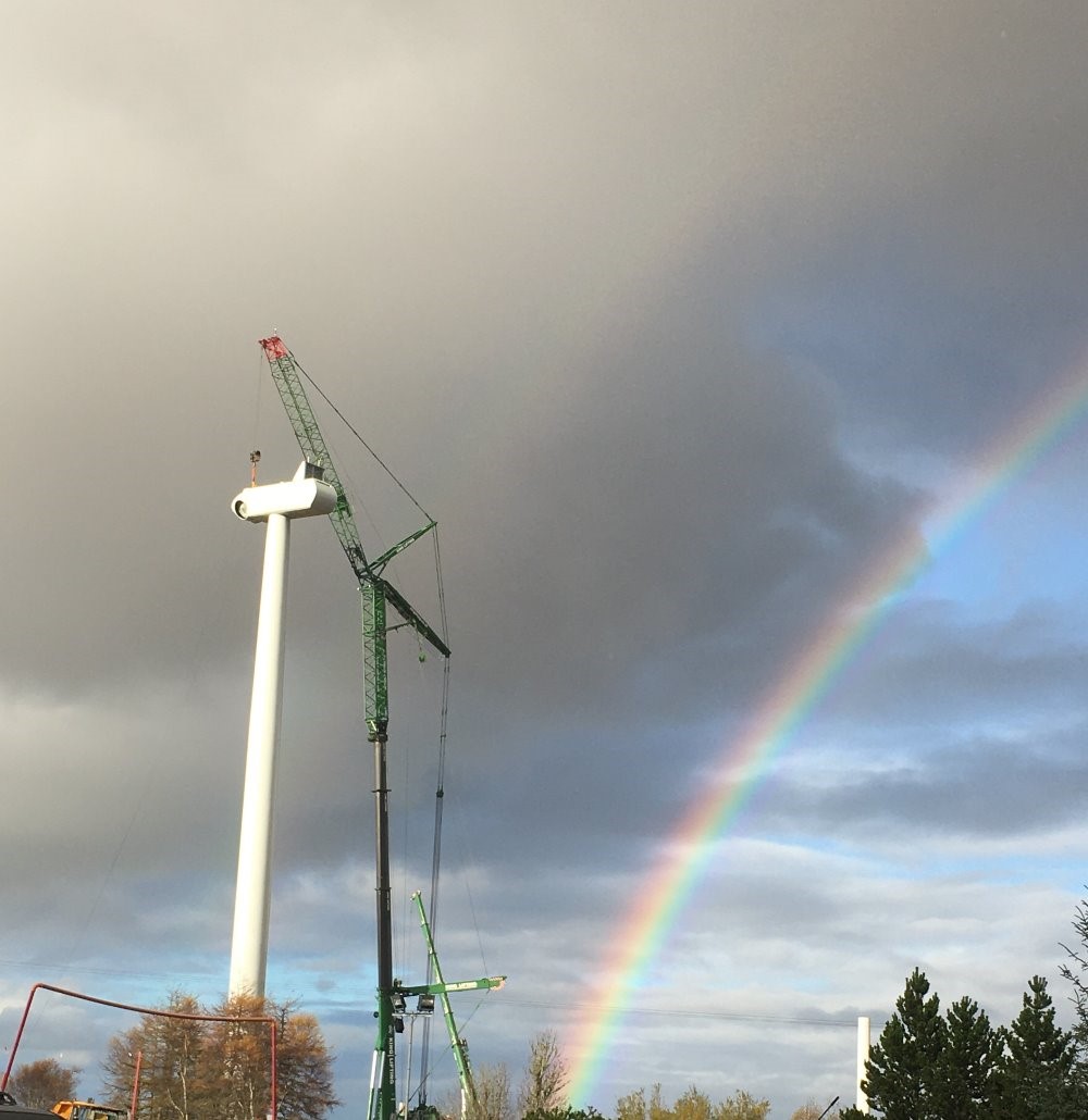 A windfarm turbine being constructed by a large crane with a bright rainbow in the background