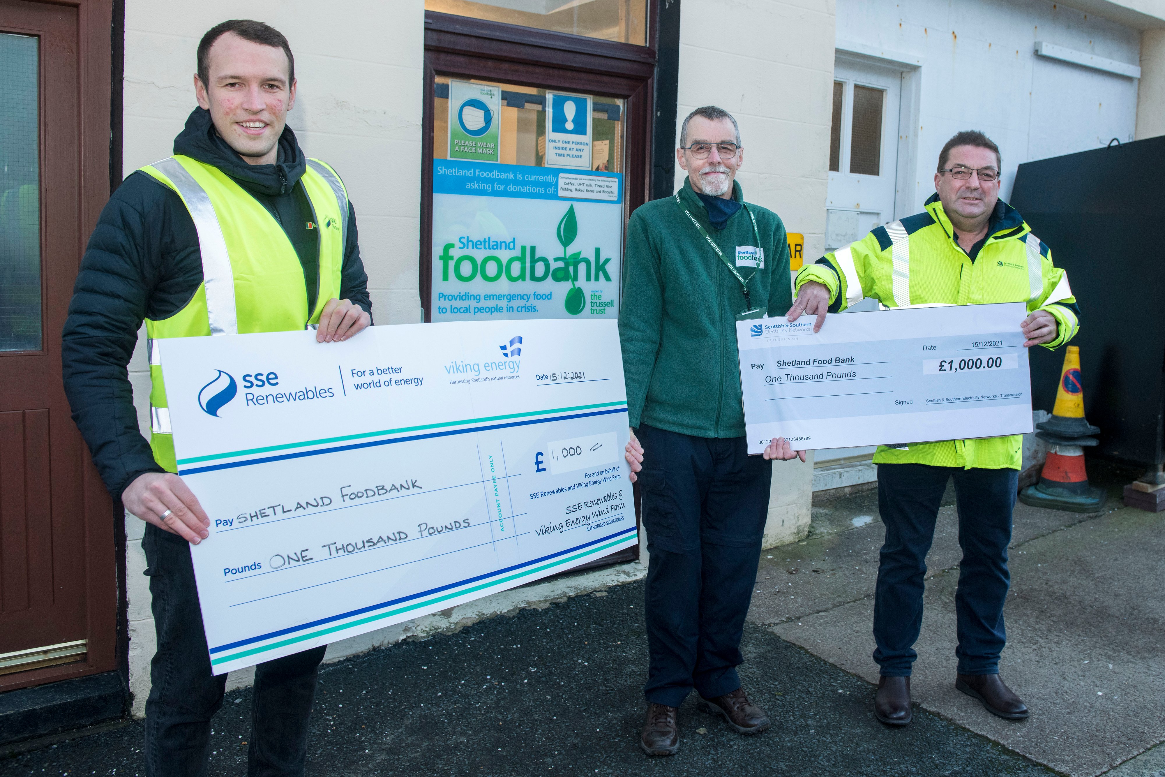 Three people standing outside Shetland Food Bank with two large novelty cheque of £1000 each 
